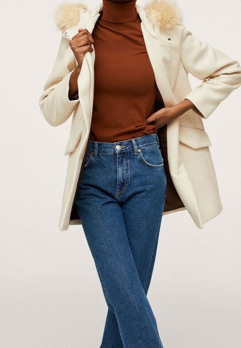 Abrigo color crema con borde de piel en la capucha, combinado con un suéter marrón de cuello alto y jeans acampanados azules. Detalles notables en los bolsillos y el acolchado.