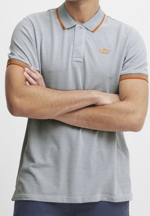 Homme portant un polo gris avec une bordure orange et les bras croisés, visible du cou à la taille sur un fond uni.