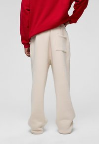 Beige sweatpants met een relaxte pasvorm en een elastische tailleband, met een enkele achterzak. Gekleed met een rode sweatshirt erboven.