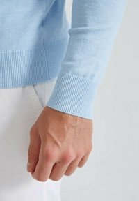 Pull bleu clair en matériau doux, avec des poignets et un bas côtelés, accompagné d'une vue rapprochée d'une main tenant le tissu.