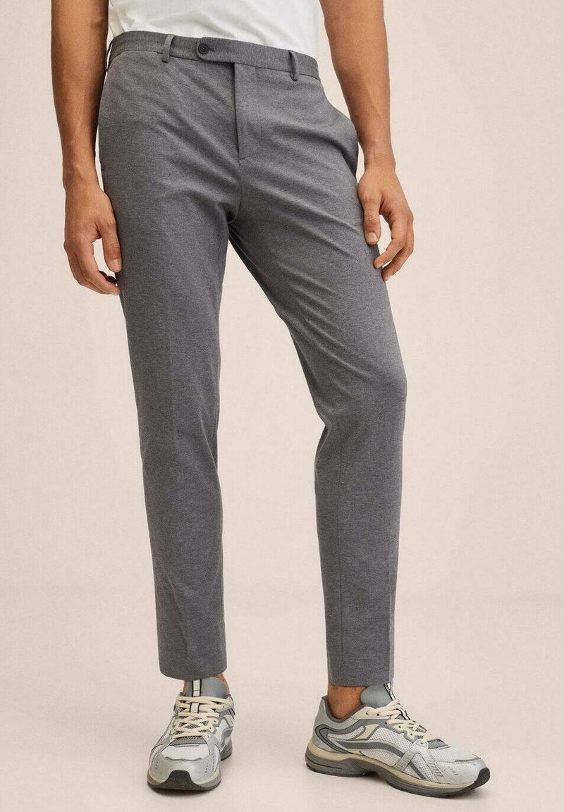 Man in slim-fit grijze broeken en grijze sport sneakers staand tegen een effen beige achtergrond.