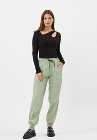 Zwarte lange mouwen shirt met een uitsnijdetail, gecombineerd met lichtgroene jogger met elastische taille. Zwarte instap-schoenen maken de outfit compleet.