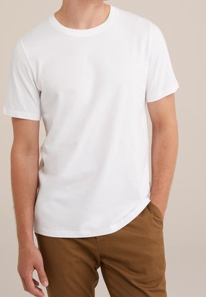 Mężczyzna w prostej białej koszulce z krótkim rękawem i brązowych spodniach, z jedną ręką w kieszeni, na neutralnym tle.