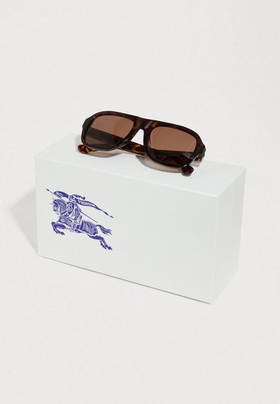 Γυαλιά ηλίου με καφέ φακούς και σχέδιο χελωνόκερου, τοποθετημένα πάνω σε λευκό κουτί που φέρει ένα μπλε ιππικό λογότυπο. Το κουτί έχει ομαλή υφή.