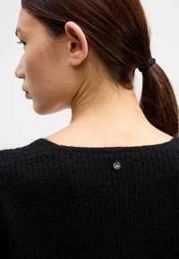 Pulover negru din tricot cu un decolteu larg, având un detaliu cu logo metalic în spate. Material texturat și croială lejeră.