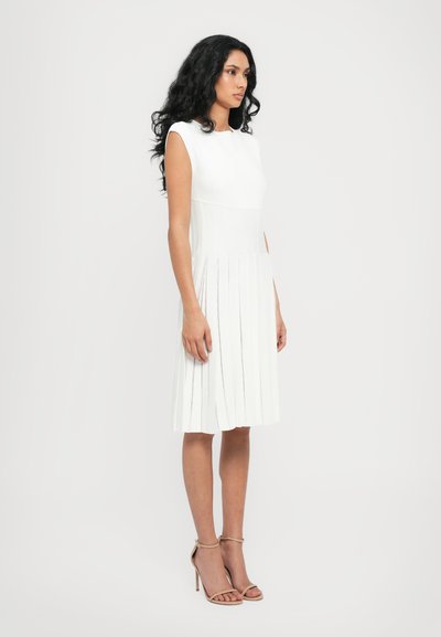 White dress with pleated skirt, cap sleeves, and fitted bodice. Smooth texture with vertical lines. Paired with strappy nude heels.
