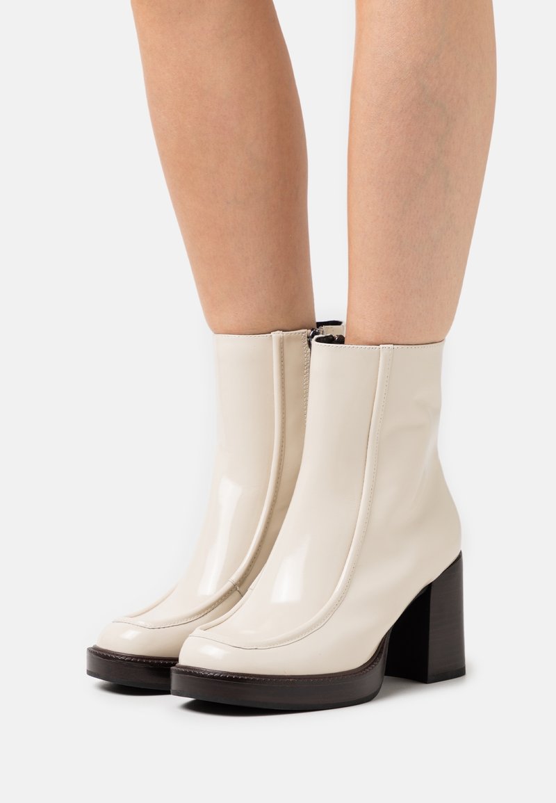Bottines beige brillantes à talons carrés portées sur des jambes nues sur un fond blanc.