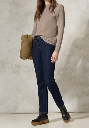 Beige textuur trui, donkerblauwe jeans met stiksel details, en zwarte enkellaarsjes met een dikke bruine zool. Houdt een olijfgroen jack vast.