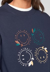 Marinblå sweatshirt med ribbad krage, prydd med fyra smiley face-grafik i koppar, grön, vit och guld, accentuerad av bladdesigner.