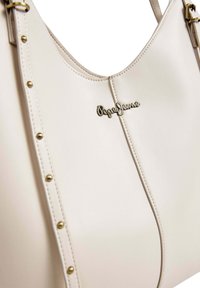 Crèmekleurige synthetische handtas met hardware in goudkleur, een logo-accent en decoratieve studs langs de zijnaad. Gladde textuur.