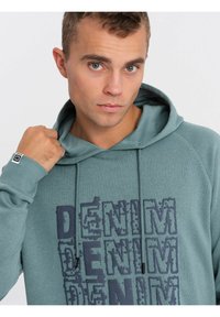 Sudadera teal hecha de tela suave, con un estampado texturizado de "DENIM" en azul oscuro. Incluye capucha con cordón y puños acanalados.