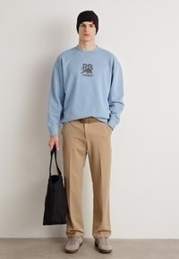 Sudadera azul claro con logotipo impreso oscuro, pantalones beiges, zapatillas grises y una bolsa de tote negra. Modelo lleva un gorro negro.