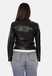 Giubbotto di pelle nera con un design aderente, polsini con zip e vita cintata. Indossato sopra jeans azzurri chiari, mostrato da dietro.