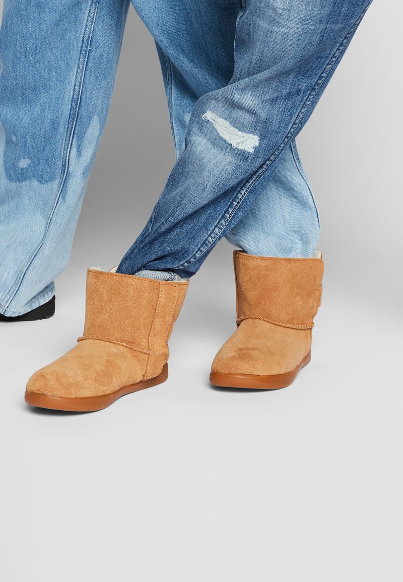 Bottes en daim beige à la cheville avec une texture douce, un bout arrondi et une semelle en caoutchouc plate, associées à un jean bleu clair déchiré.
