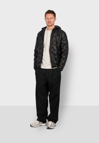 Sort pufferjakke med en quiltet mønster, hætte og lynlåslukning, parret med en hvid t-shirt og løstsiddende sorte bukser. Grå sneakers fuldender looks.