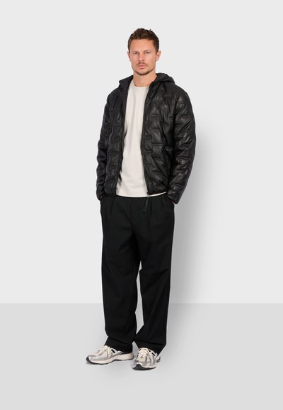 Doudoune noire avec un motif matelassé, capuche et fermeture éclair, associée à un t-shirt blanc et un pantalon noir ample. Des baskets grises complètent le look.