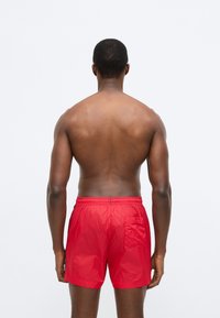 Piros úszónadrág könnyű, fényes anyagból. Rugalmas derékrésszel és hátsó zsebbel rendelkezik. A szabás bő, a hossz pedig a comb közepéig ér.