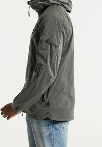 Szara kurtka z kapturem o teksturowanym wykończeniu, z kieszeniami na zamek i guzikiem na lewym rękawie. Noszona z jasnoniebieskimi dżinsami.