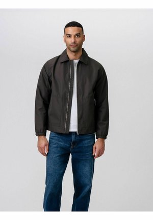 Férfi fekete cipzáras dzsekiben, fehér inggel és kék farmerral, sima világosszürke háttér előtt állva.