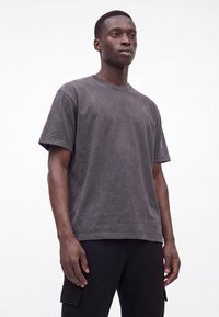 Homme portant un t-shirt uni gris foncé à manches courtes et un pantalon noir, debout devant un fond blanc.
