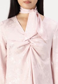 Blusa in raso rosa con motivo floreale, dettagli con nodo al collo, e tessuto arricciato in vita, con maniche lunghe e una texture liscia.