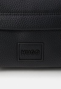 Borsa nera in pelle con finitura testurizzata, bordi arrotondati e logo "HUGO" impresso sul fronte. Fodera interna liscia.