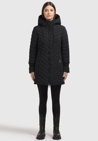 Kobieta ubrana w czarną, długą pikowaną kurtkę z kapturem, zapinaną na zamek, czarne legginsy i czarne botki, stojąca na białym tle.