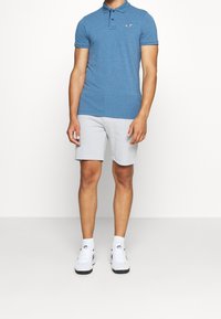 Modrá polo košile s krátkým rukávem, s knoflíkovou légou a logem; spárovaná se světle šedými šortkami a bílými sportovními botami. Jednoduché pozadí.