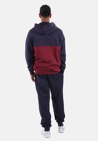 Completo con felpa con cappuccio e pantaloni in blocchi di colore blu navy e bordeaux, realizzato in tessuto morbido, con cappuccio regolabile con coulisse e polsini elasticizzati.