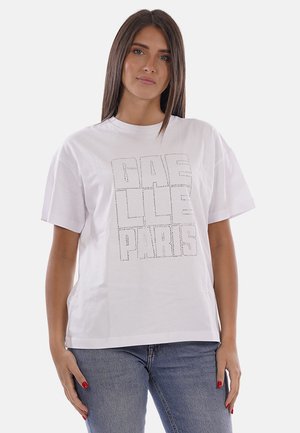 Donna con capelli castani lisci che indossa una t-shirt bianca oversize con la scritta "GAS LLS PARIS" e jeans blu, in piedi davanti a uno sfondo semplice.