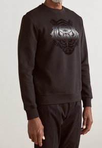 Sudadera negra con un diseño texturizado de cabeza de tigre en gris en el frente. Fabricada con tejido suave, tiene cuello redondo y puños acanalados.
