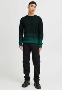 Donkergroene gebreide trui met een gestructureerd patroon, effen groene band aan de onderkant, gecombineerd met zwarte jeans en zwarte veterschoenen.