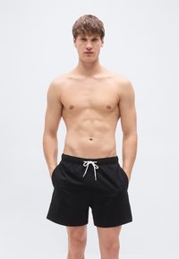 Μαύρα μαγιό σορτς με υφή, ελαστική μέση και λευκό κορδόνι. Το μοντέλο στέκεται με τα χέρια στις τσέπες μπροστά σε απλό φόντο.