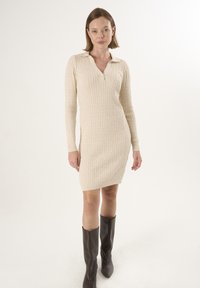 Abito lavorato a maglia beige con scollo a V e colletto, caratterizzato da un motivo a treccia e maniche lunghe, abbinato a stivaletti marroni al ginocchio.