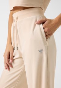 Beige Sporthosen mit weicher Textur, ausgestattet mit einem Kordelzugbund, Seitentasche und einem dekorativen dreieckigen silbernen Akzent.