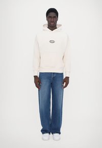 Kräms hoodie med svart logotyp, känguruficka; blå vida jeans; vita sneakers; enfotad bakgrund.