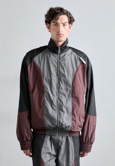 Veste en windbreaker bicolore en gris, noir et bordeaux. Dotée d'un col haut et d'une fermeture éclair, avec des poignets et une taille élastiques.