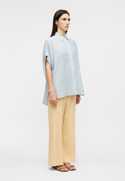 Femme debout portant une chemise à manches courtes ample à rayures bleu clair, un pantalon beige à jambes larges et des sandales marron, sur un fond uni.
