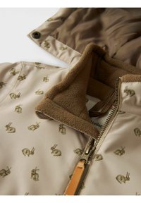 Chaqueta beige con estampado de conejos, cuello de forro polar marrón suave y cierre con cremallera. Tejido exterior liso, interior forrado con material marrón.