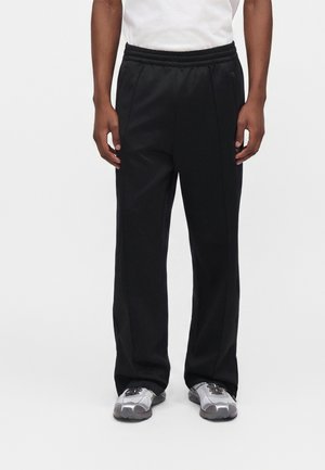 Man draagt losse zwarte broek met elastische tailleband en grijze sportschoenen, staand tegen een eenvoudige lichte achtergrond.