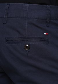 Dettaglio ravvicinato della tasca posteriore di pantaloni blu navy con chiusura a bottone e piccolo logo rettangolare rosso, bianco e blu sopra la tasca.