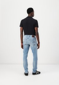 Moški v črnem polo majici, svetlih modrih kavbojkah in črnih supergah z belimi črtami, stoji obrnjen stran od fotoaparata proti enobarvni beli ozadju.