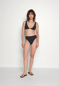 Černé bikiny s trojúhelníkovým horním dílem a kalhotkami s vysokým pasem, vyrobené z hladké látky; modelka stojí v žabkových sandálech, jednoduché pozadí.