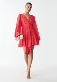 Rotes Wickelkleid mit langen Ärmeln, strukturiertem Stoff, asymmetrischem Saum und geraffter Taille. Das Model hält eine glänzende weiße Clutch und trägt goldene Absätze.