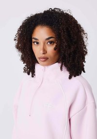 Lichtroze ritssweater met hoge kraag, witte accentstiksels en logo. Zachte textuur, relaxed fit en lange mouwen.