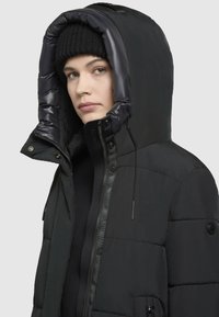 Schwarze Steppjacke mit Kapuze, ausgestattet mit einer glatten Oberfläche, einem Reißverschluss und einer strukturierten, dunklen Strickmütze darunter.