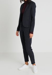 Granatowy garnitur w paski z dopasowanym żakietem i dopasowanymi spodniami, zestawiony z bordowym golfem i białymi tenisówkami.