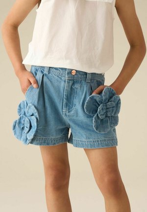 Barn iført blå denimshorts med store stofblomster på hver lomme og en hvid ærmeløs top.