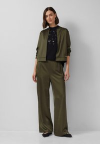 Olivegrüner Bomberjacke und passende weit geschnittene Hosen aus glattem Stoff, kombiniert mit einem schwarzen Oberteil mit Paillettenverzierungen. Schwarze Schuhe runden den Look ab.
