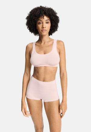 Vrouw met krullend haar die een lichtroze sportbeha en bijpassende shorts met hoge taille draagt, staand tegen een effen achtergrond.